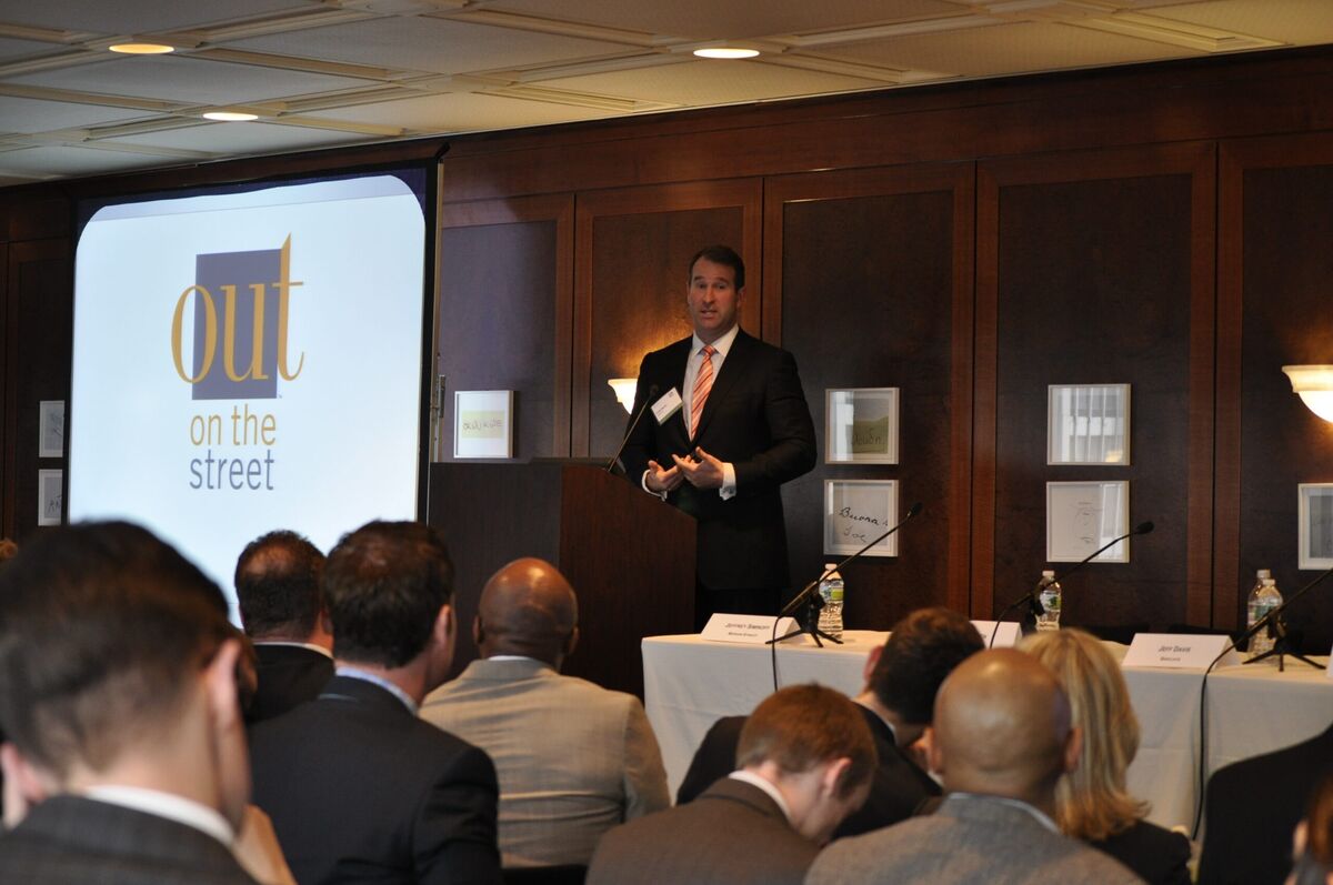 A speaker at a podium addresses a packed room at the first Out on the Street summit, with the Out on the Street logo on screen